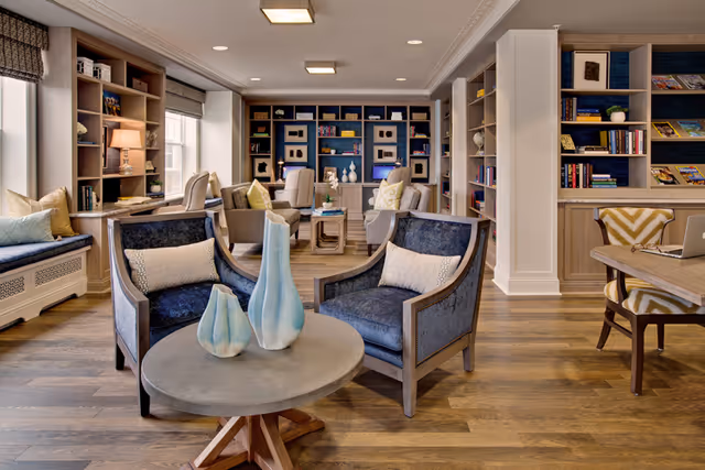A cozy and well-lit living room area with wooden flooring, featuring two blue upholstered armchairs with cushions around a round table holding three decorative vases. The room has built-in bookshelves filled with books and decorative items, a window seat with cushions, and additional seating including armchairs and a desk with a chair. The overall decor is warm and inviting with neutral tones and soft lighting.