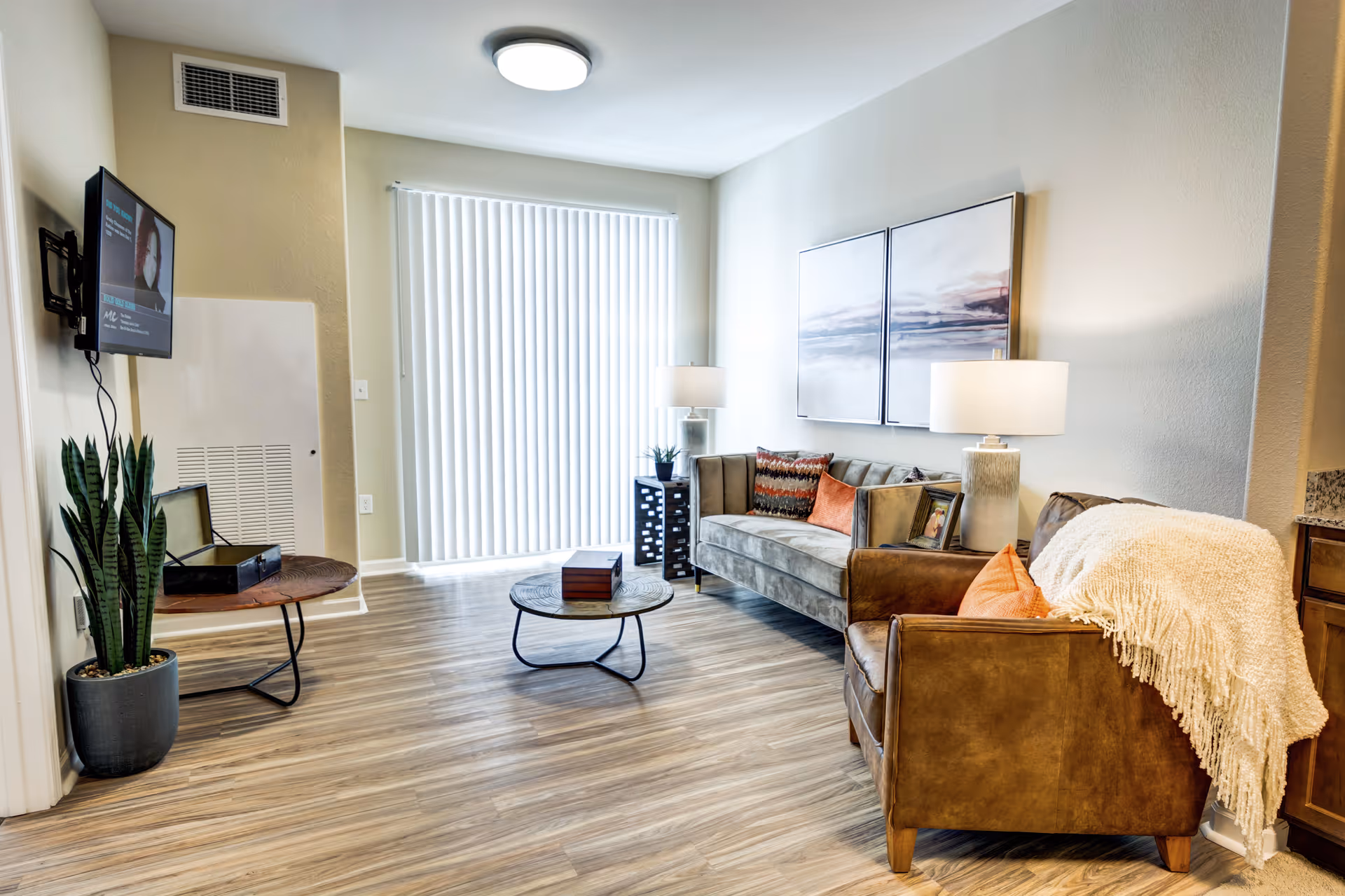A bright and modern living room in a senior living facility featuring a gray sofa with colorful pillows, a brown leather armchair with a white throw blanket, a round coffee table, a side table with a lamp, a potted plant, and a wall-mounted TV. The room has wood flooring and vertical blinds covering a large window.