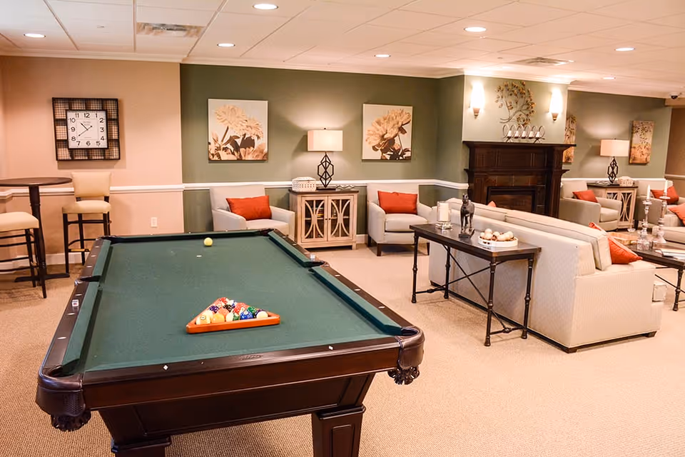 A cozy common area with a pool table in the foreground, surrounded by comfortable seating including armchairs and sofas with red cushions. The room features warm lighting, wall art with floral designs, a clock on the wall, and a decorative fireplace in the background.