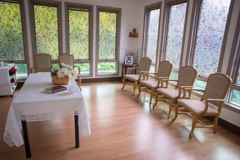 Bright sitting room with a table covered in a white cloth and a basket of flowers, and a row of wooden armchairs by tall windows with patterned shades.