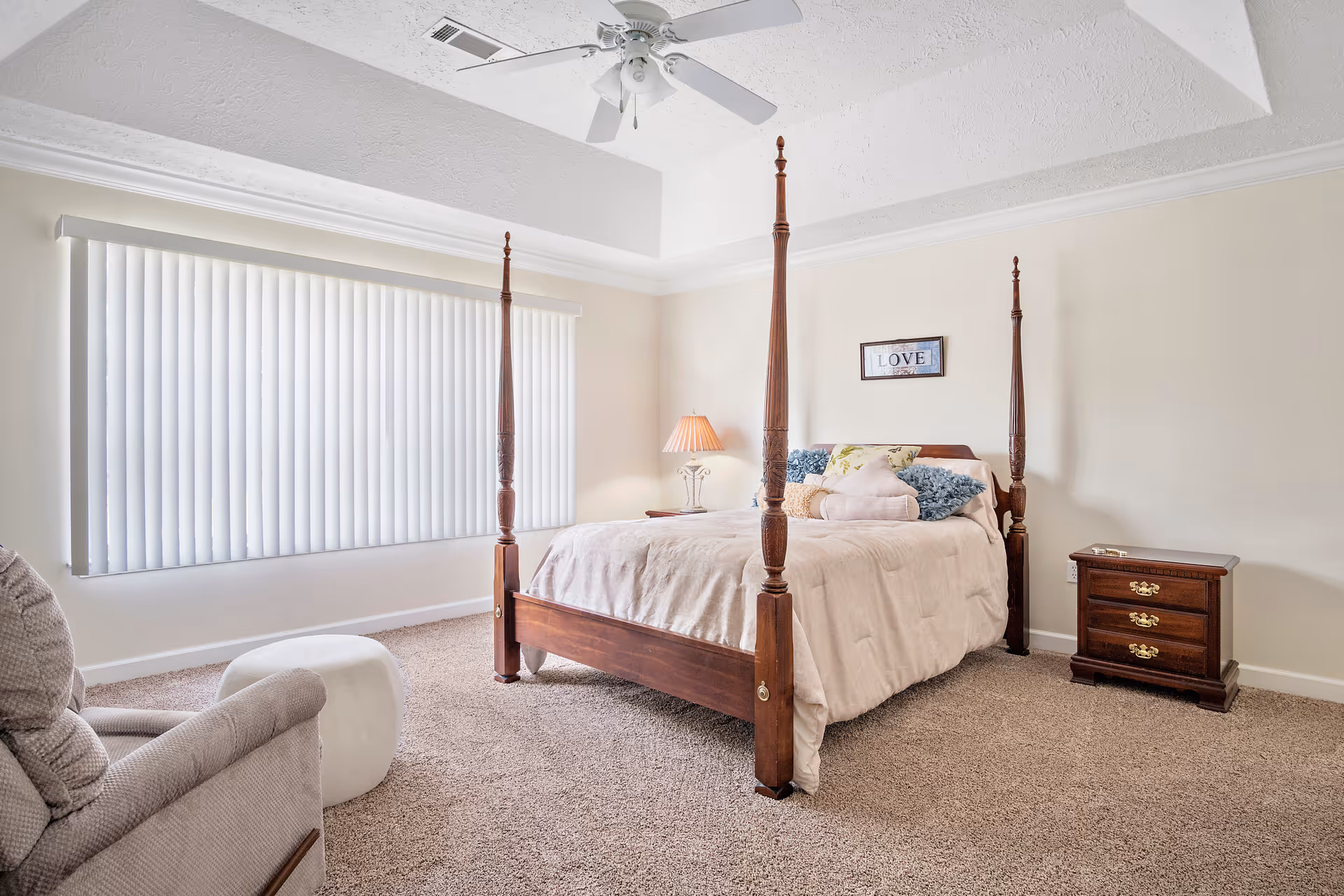 A bright bedroom with a four-poster wooden bed covered with a beige comforter and multiple pillows. There is a wooden nightstand with a lamp on the right side of the bed and a framed picture with the word 'LOVE' hanging on the wall above the bed. A beige recliner chair and a white ottoman are positioned near a large window with vertical blinds. The ceiling has a white fan and the floor is carpeted.