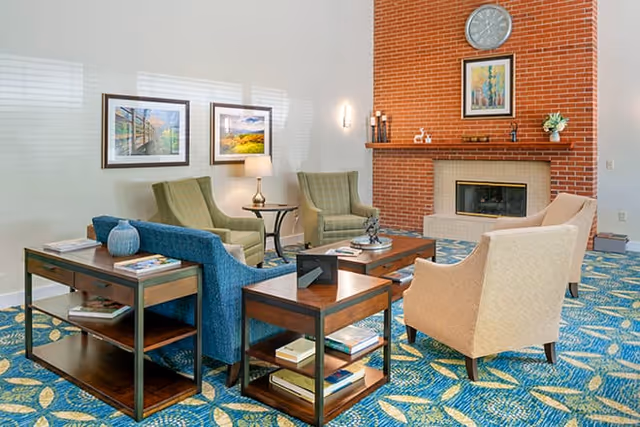 A cozy living room area with a blue patterned carpet, two beige armchairs, two green armchairs, and a blue sofa arranged around two wooden coffee tables. A brick fireplace with a clock and framed artwork above it is on the far wall. There are two framed pictures on the adjacent wall and a small table lamp between the green armchairs.