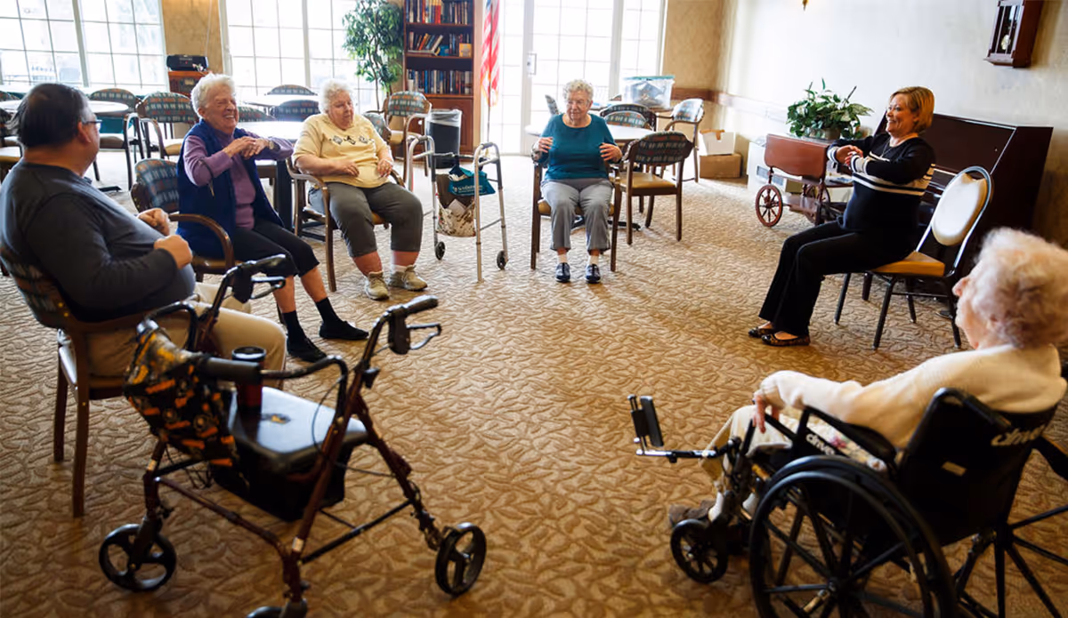A group of elderly people sitting in a circle in a carpeted room, engaging in a seated exercise or activity led by a woman. Some participants use walkers or wheelchairs. The room has large windows, bookshelves, and plants.