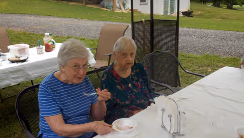 Two elderly women sitting at a table outdoors with white tablecloths, one woman wearing a blue striped shirt eating from a bowl, and the other woman wearing a floral dress. There are chairs around the table and a grassy area with a gravel path in the background.