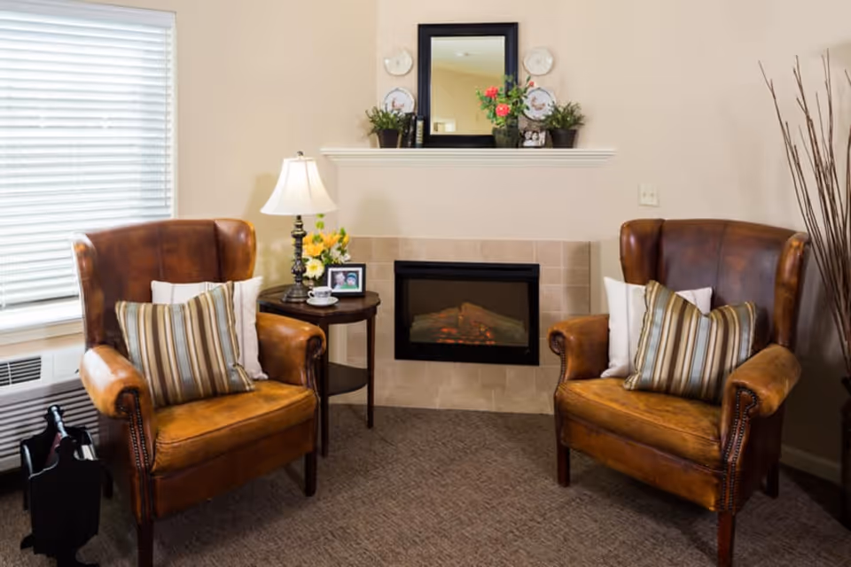Two leather armchairs flank a small side table and an electric fireplace with a mirror and decor above it in a cozy living room seating area.