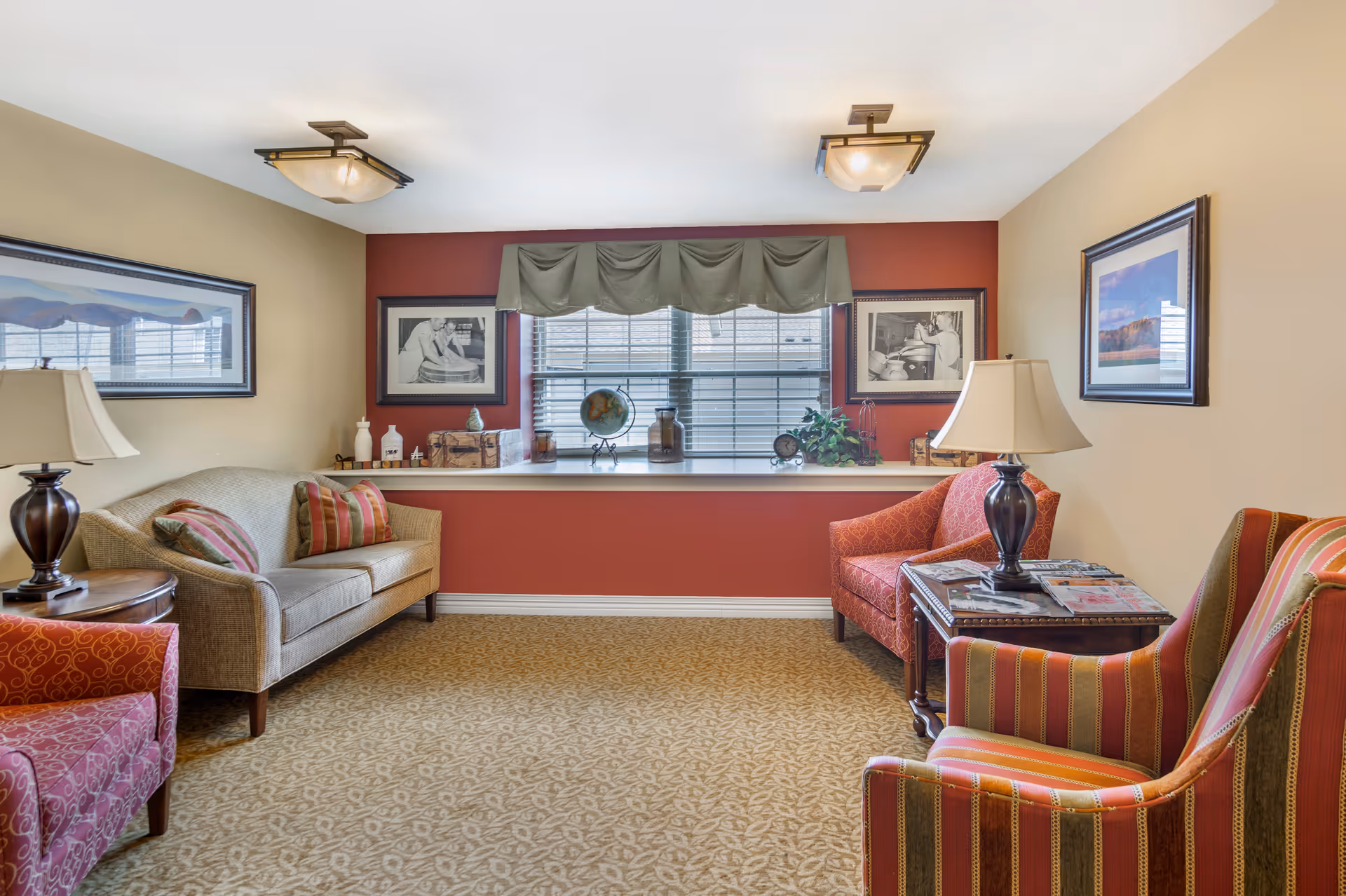 A cozy living room area with a beige sofa and three patterned armchairs arranged around two wooden side tables with lamps. The walls are painted beige and red, decorated with framed pictures. A large window with blinds and a green valance lets in natural light, and the windowsill holds decorative items including a globe, jars, and a small clock. The floor is carpeted with a patterned design.