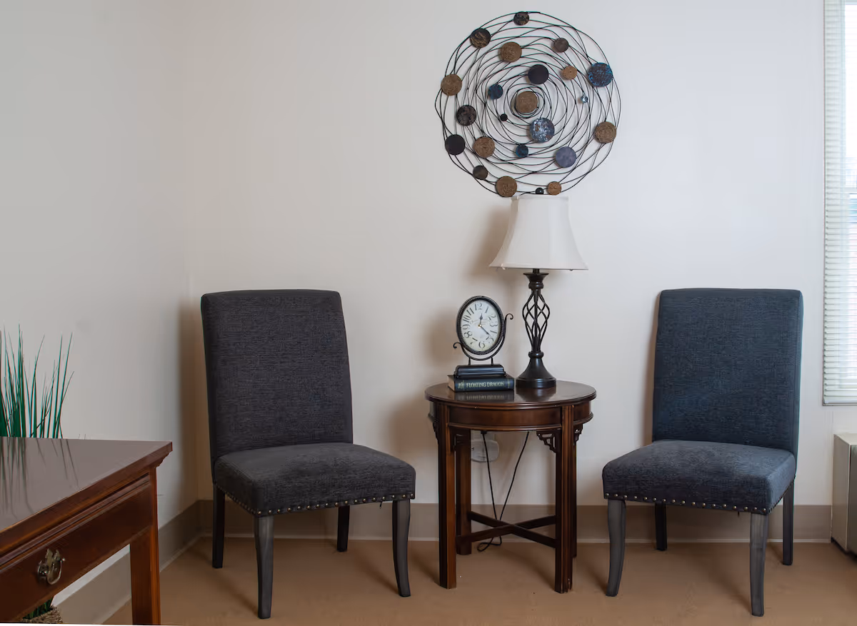 A cozy sitting area with two dark gray upholstered chairs placed on either side of a round wooden side table. On the table, there is a decorative clock and a lamp with a white shade. Above the table, a circular metal wall art piece with various round elements is mounted on a plain white wall. To the left, part of a wooden desk is visible, and to the right, a window with blinds is partially shown.