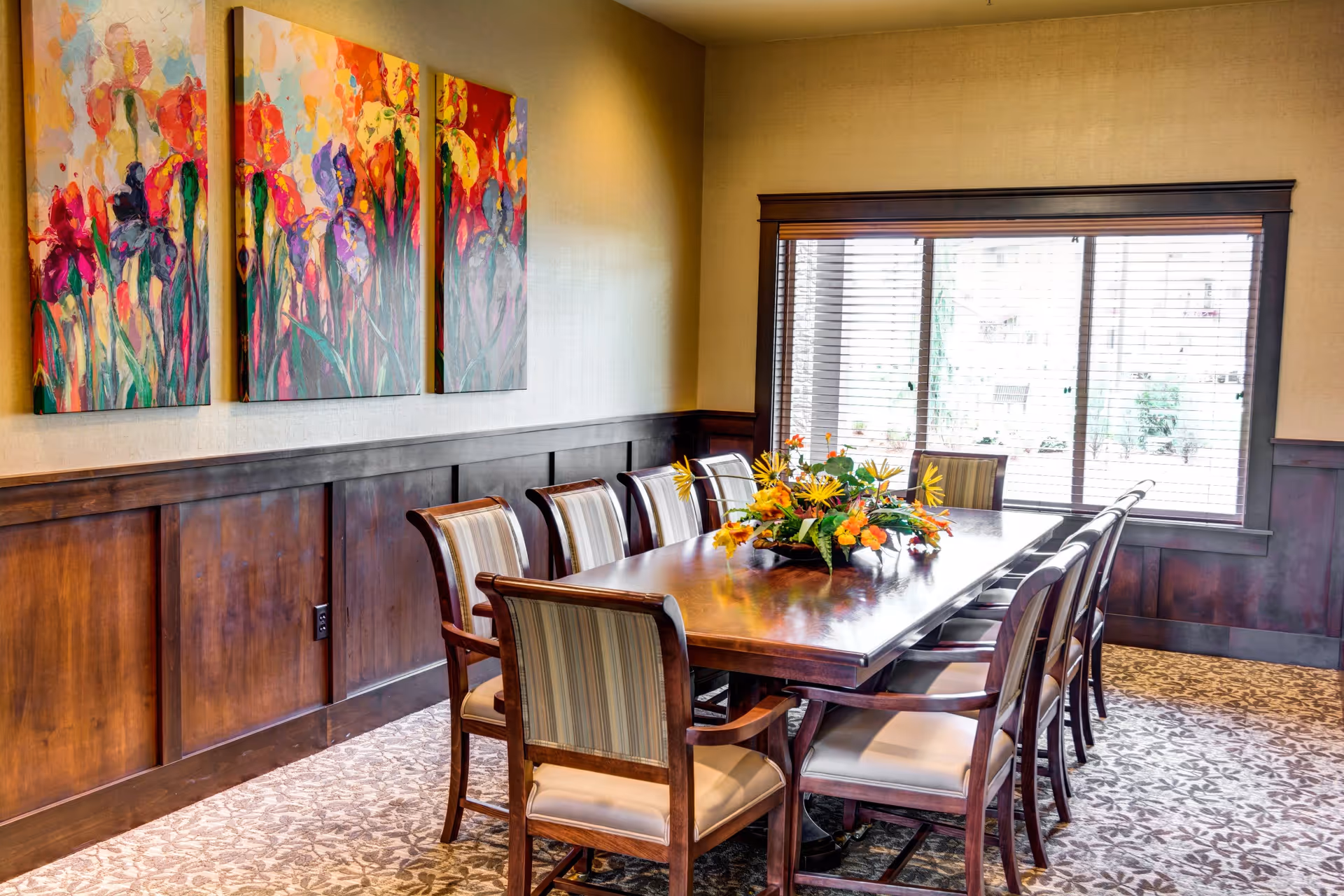 A well-lit dining room with a long wooden table surrounded by ten cushioned chairs. A floral centerpiece decorates the middle of the table. The room features wood paneling on the lower half of the walls, a large window with blinds, and three colorful abstract paintings on the wall.