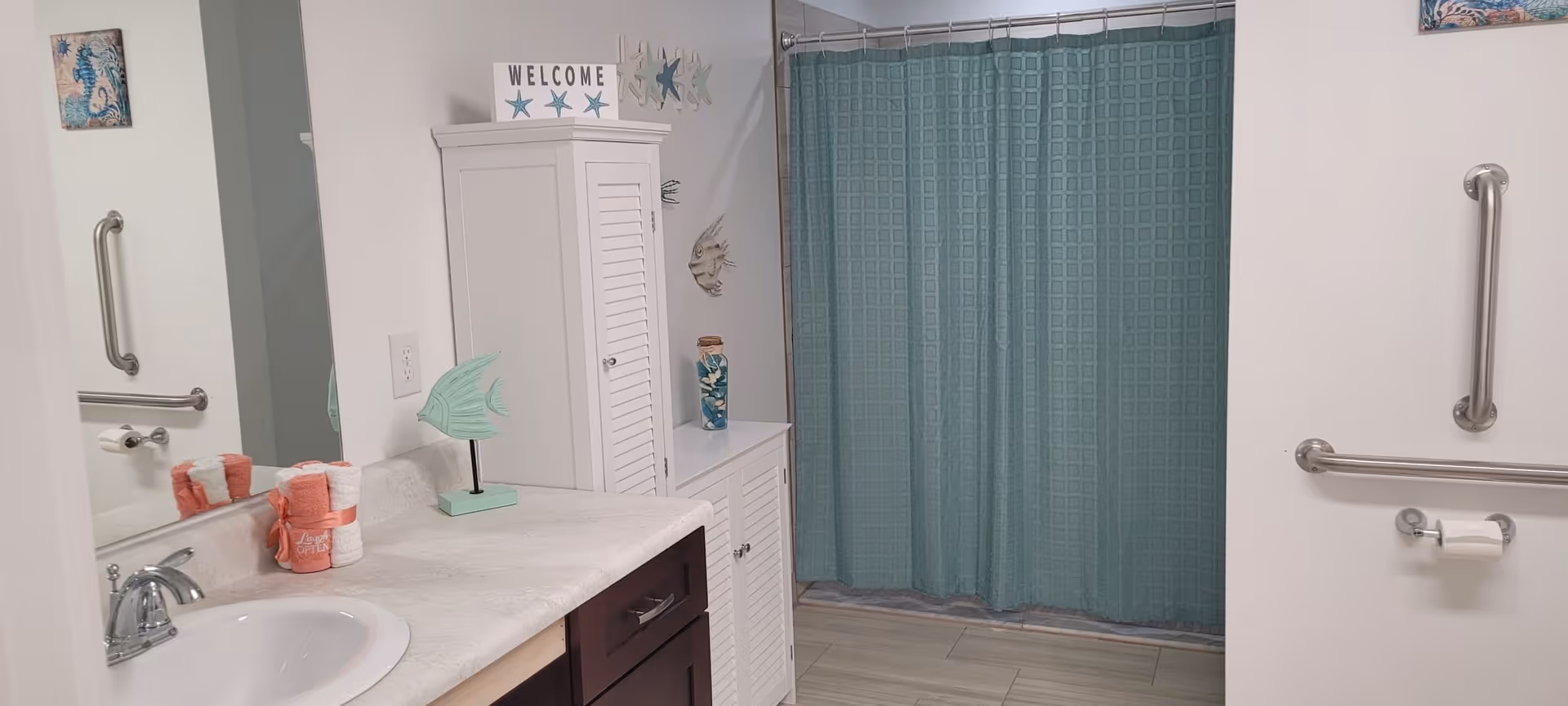 A bathroom with a white sink and countertop, dark brown cabinets, a white storage cabinet, and a teal shower curtain. The walls are white with silver grab bars and a toilet paper holder. Decorative items include a small green fish sculpture, a 'WELCOME' sign with starfish, and a jar with seashells. There are also rolled coral-colored towels on the countertop.
