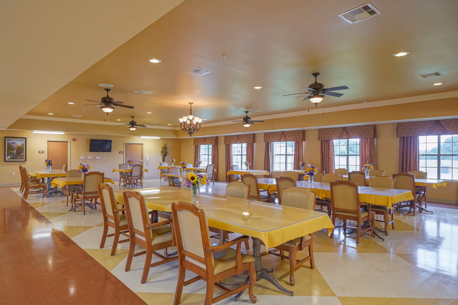 Spacious senior living dining room with multiple tables covered in yellow tablecloths, chairs, vases of flowers, ceiling fans, and large windows.