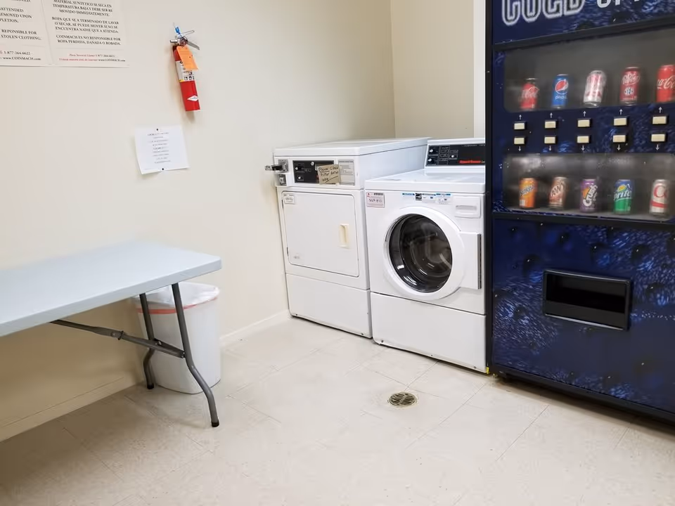 A laundry room with a white washing machine and dryer side by side, a blue vending machine stocked with various soda cans, a folding table, a trash can, and a fire extinguisher mounted on the wall. There are also some notices posted on the wall above the fire extinguisher.
