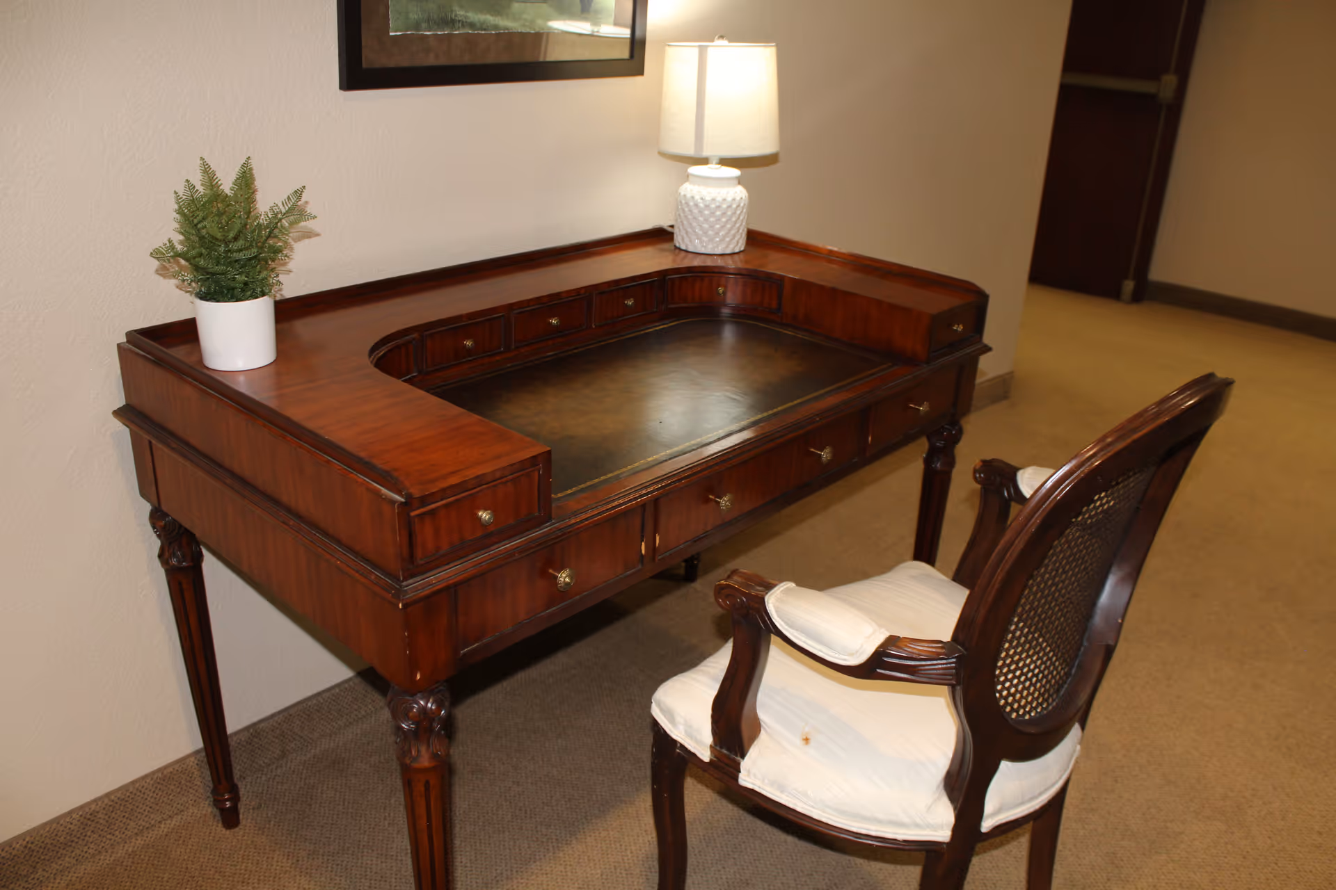 Wooden writing desk with a lamp, potted plant, and an upholstered chair in a carpeted interior hallway.
