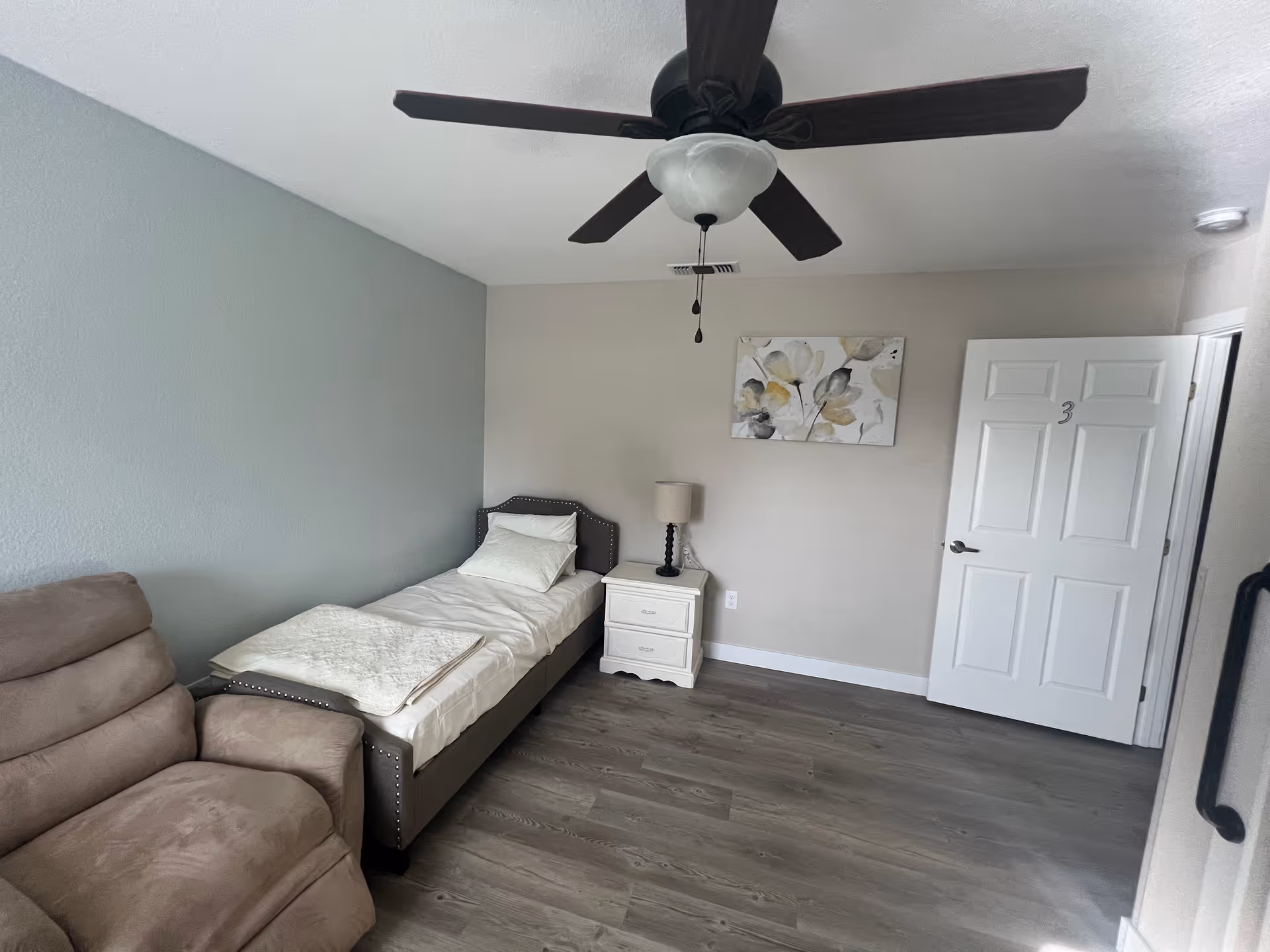 A simple bedroom with a single bed, nightstand and lamp, a recliner, ceiling fan, and a door marked "3".