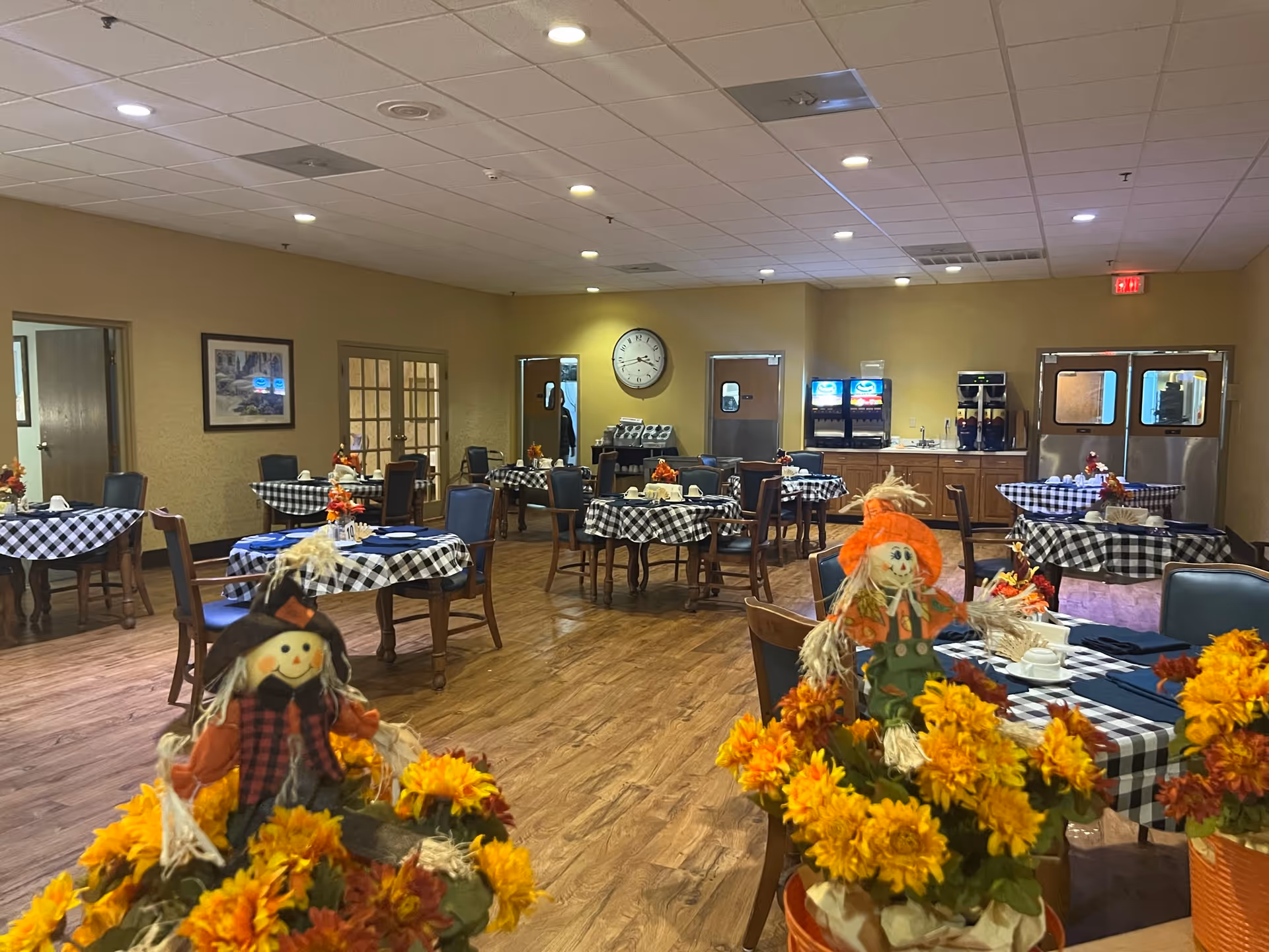 A senior living facility dining room with multiple round tables covered in black and white checkered tablecloths. Each table is set with cups and napkins, and decorated with autumn-themed centerpieces including small scarecrow figures and yellow flowers. The room has wood flooring, beige walls, and a ceiling with recessed lighting. In the background, there are beverage dispensers and double doors.
