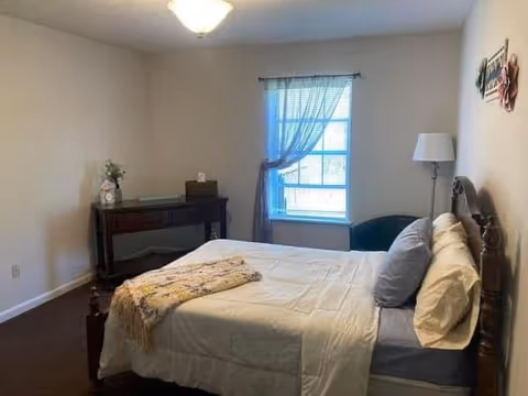 A bedroom with a neatly made bed featuring white and gray bedding and a decorative throw. There is a wooden headboard, a floor lamp beside the bed, a window with sheer curtains, and a dark wooden console table with decorative items against the wall.