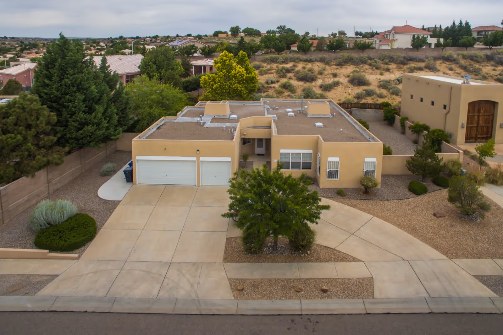 Aerial view of a single-story beige building with a flat roof and a two-car garage, surrounded by a desert landscape with sparse vegetation and neighboring houses in the background.