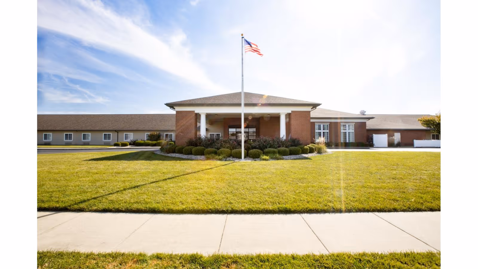 Front exterior of a single-story brick senior living building with a central portico, American flag on a flagpole, and a manicured lawn.