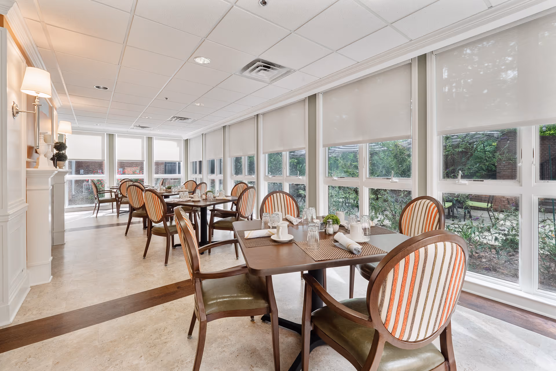 Bright dining room with multiple tables and chairs arranged neatly. Each table is set with placemats, napkins, glasses, and small decorative plants. Large windows with white roller shades allow natural light to fill the room, and there are wall-mounted lamps on the left side.