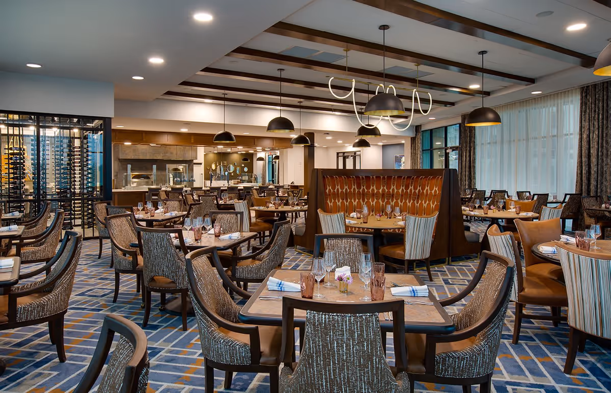 A spacious, well-lit dining room with multiple tables set with glasses, napkins, and small flower vases. The room features a patterned carpet, upholstered chairs, pendant lighting, and a wine rack on the left side. Large windows with curtains allow natural light to enter the space.