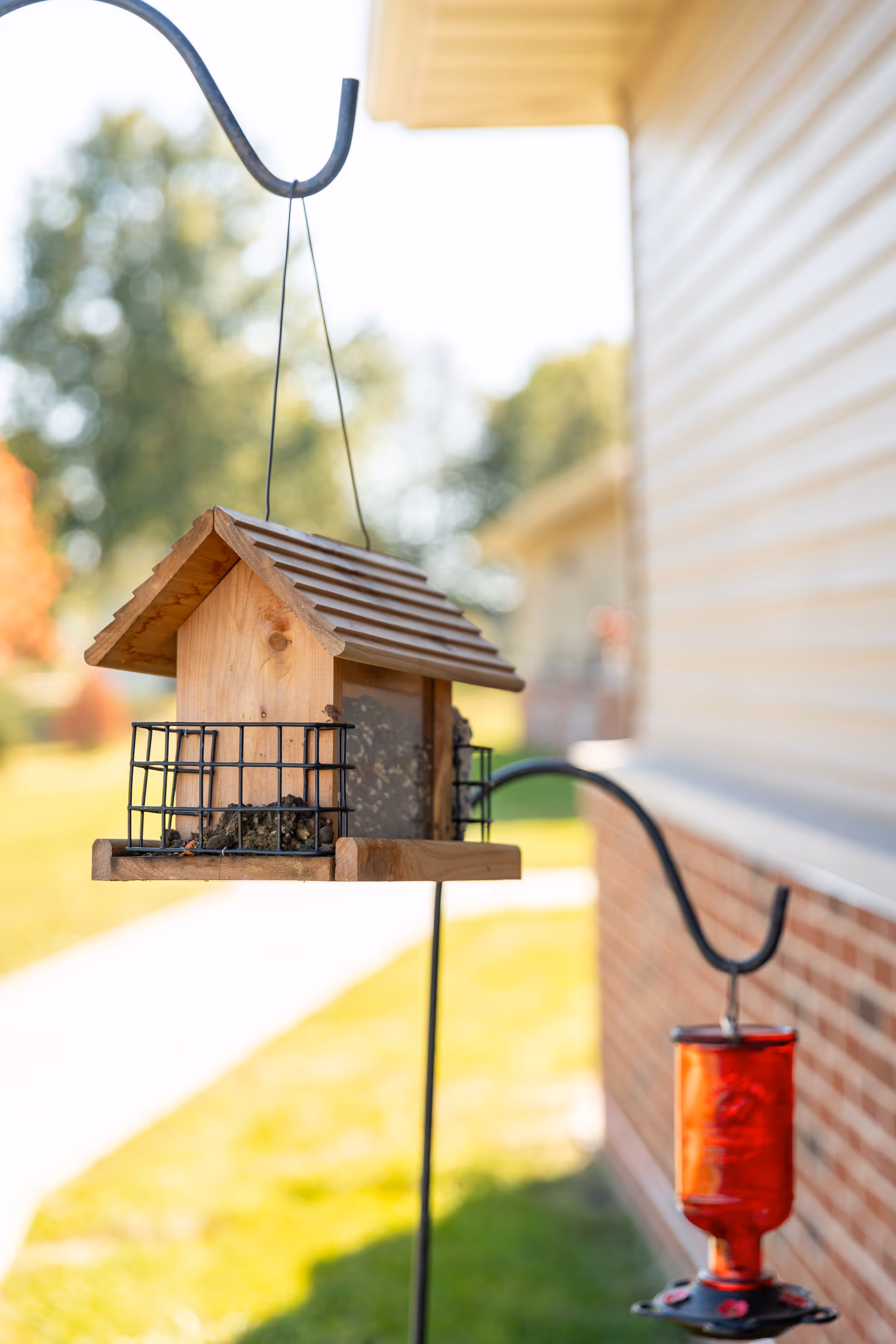 A wooden bird feeder hanging on a metal hook outside near a building with beige siding and a brick lower wall, with a red hummingbird feeder hanging on a separate hook in the background and green grass and trees blurred in the distance.
