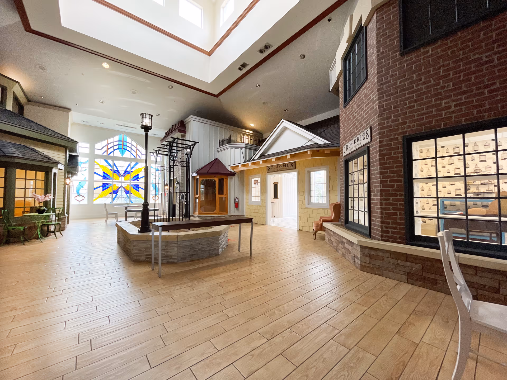 Indoor common area designed to resemble a small town street with storefronts including a grocery store and a building labeled St James. The space features wooden flooring, a central bench around a lamppost, large stained glass windows, and high ceilings with skylights.
