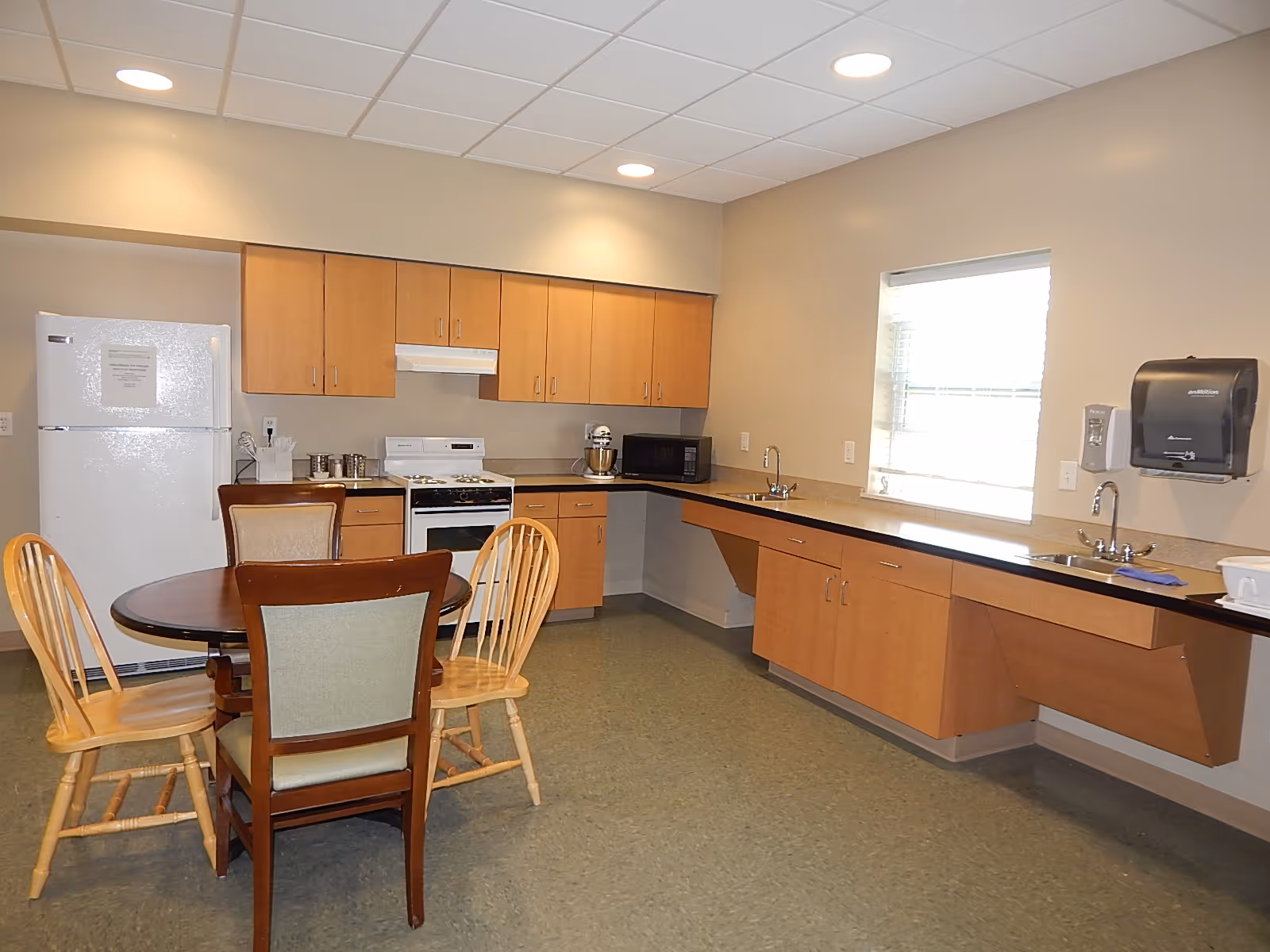 Communal kitchen and dining area with a round table and chairs, refrigerator, stove, cabinets, countertop sinks and a window.