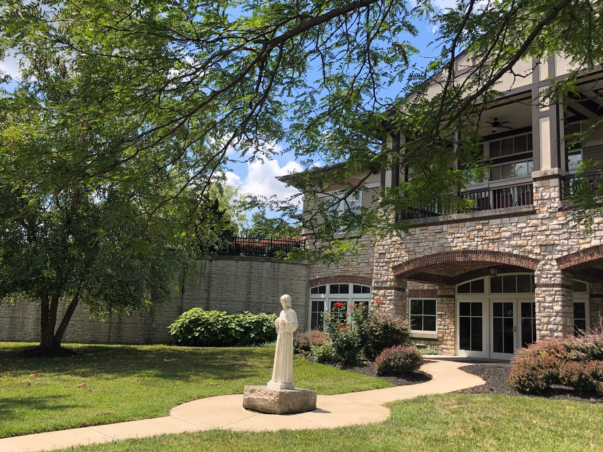 Outdoor view of a senior care facility with a stone building featuring arched doorways and a balcony. In the foreground, there is a statue of a person standing on a stone pedestal surrounded by a curved concrete pathway, green grass, bushes, and trees under a partly cloudy sky.