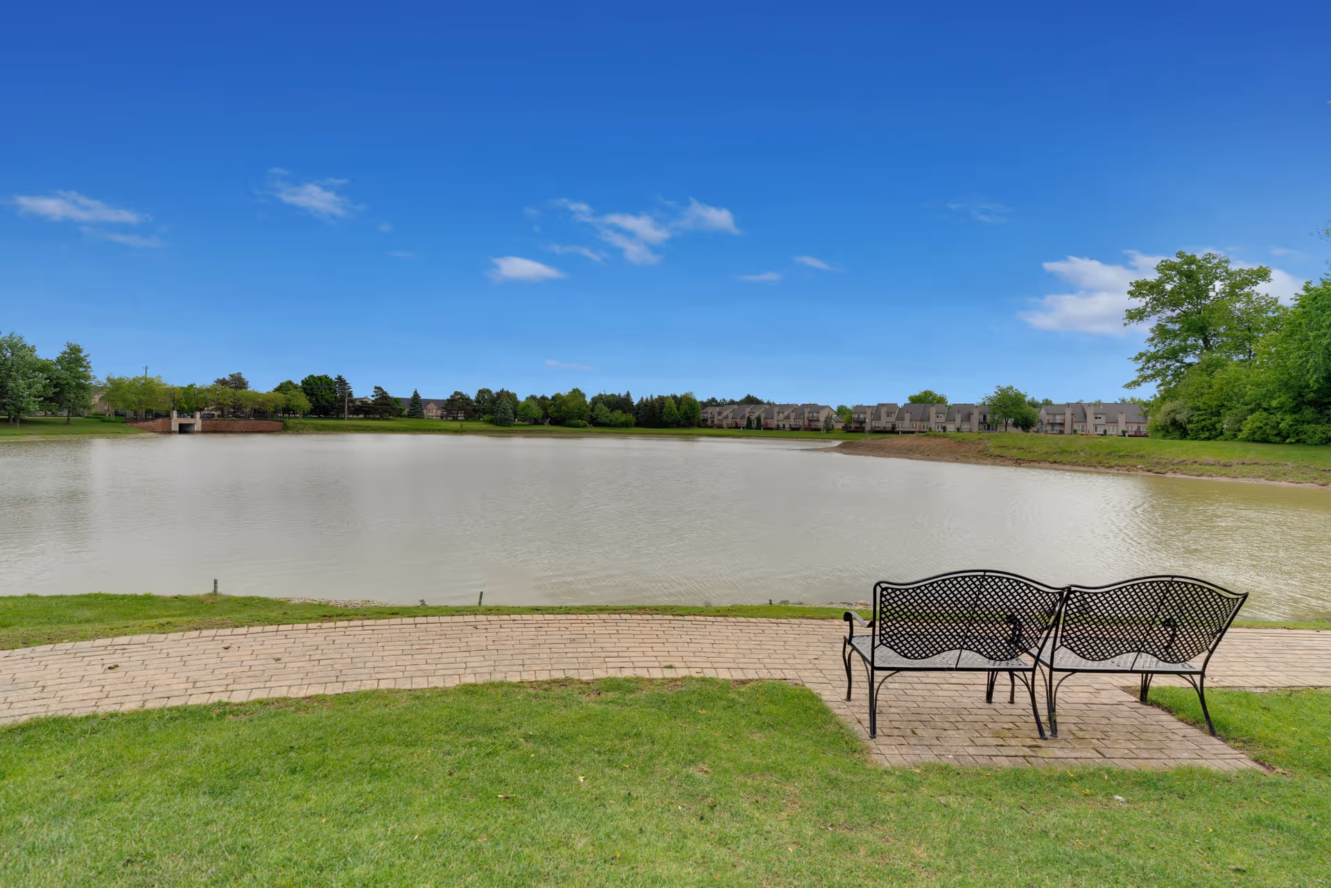 Two black metal benches on a brick patio facing a large pond with green grass and trees surrounding it under a clear blue sky.