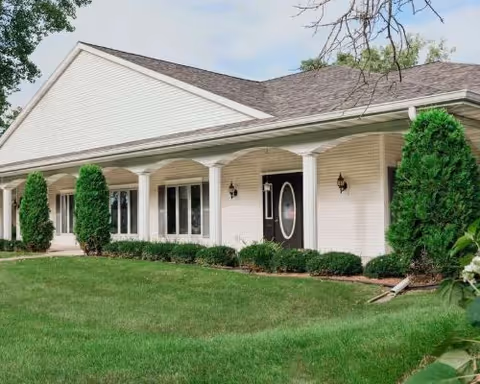 Exterior view of a single-story assisted living facility building with white siding, a covered porch supported by columns, a black front door with an oval window, and neatly trimmed bushes and green lawn in front.