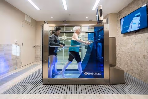 An elderly woman walking on an underwater treadmill inside a HydroWorx aquatic therapy tank, assisted by a caregiver in a well-lit therapy room with beige walls and a mounted screen displaying information.
