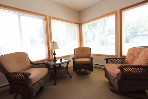 A cozy corner seating area with three wicker chairs with cushions arranged around a small wicker side table holding a lamp, set against three large windows with blinds letting in natural light.