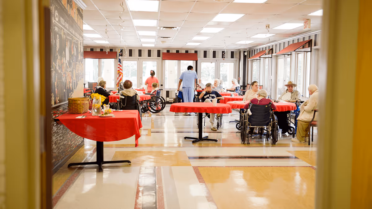 A dining room in Tallwoods Care Center with elderly residents seated at tables covered with red tablecloths. Some residents are in wheelchairs, and a caregiver in blue scrubs is assisting them. The room has large windows with red valances, striped wallpaper, and an American flag near the back wall.