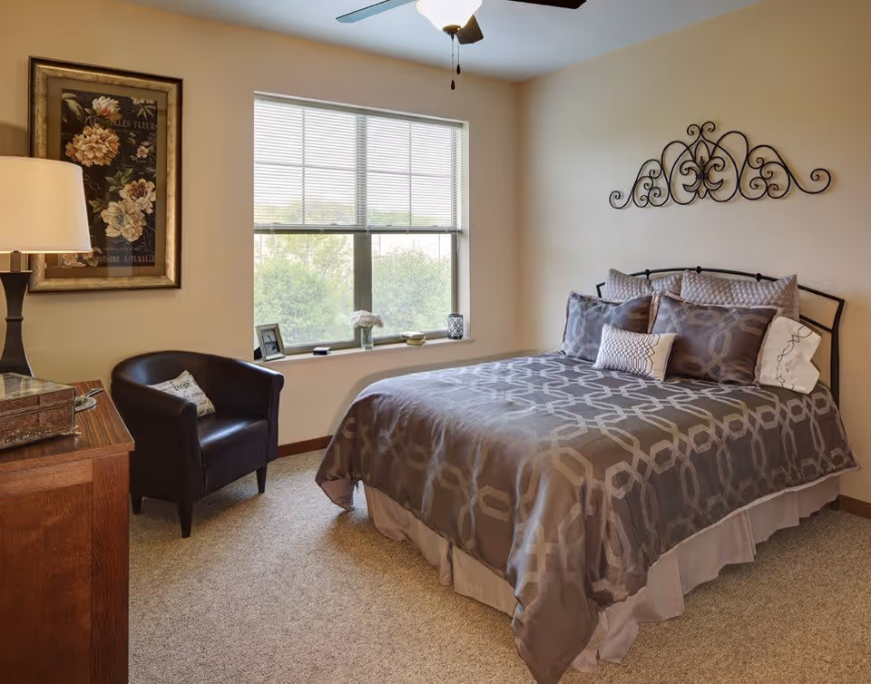 A cozy bedroom with a neatly made bed featuring a patterned gray comforter and multiple pillows. There is a black armchair with a small pillow next to a wooden dresser with a lamp and decorative box. A large window with blinds lets in natural light, and a framed floral artwork hangs on the wall beside the window. Above the bed is an ornate metal wall decoration.