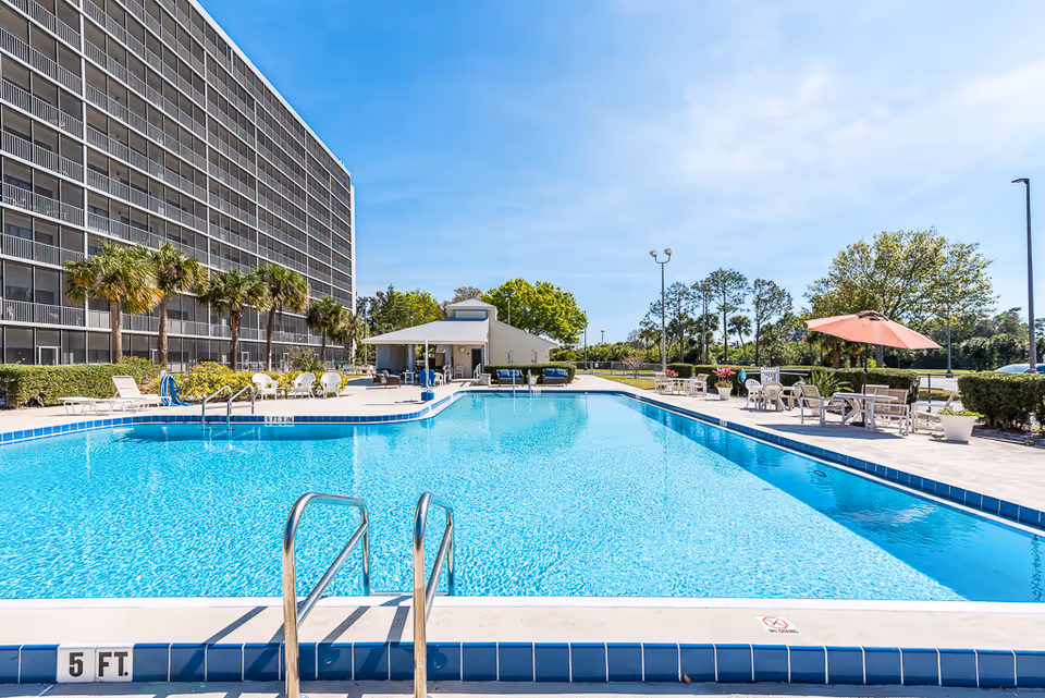 Outdoor swimming pool with lounge chairs, umbrellas, and a multi-story building in the background.