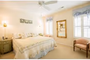 A bright and cozy bedroom with a large bed covered in a light-colored floral bedspread. The room features two windows with white shutters and blue valances, a ceiling fan with a light, a wooden chair with a cushion, a large framed mirror on the wall, and two bedside tables with lamps.