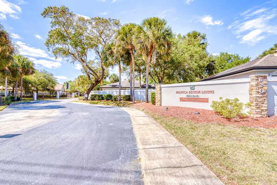 Entrance driveway and landscaped front sign reading 'Pacifica Senior Living Belleair' with palm trees and facility buildings behind.
