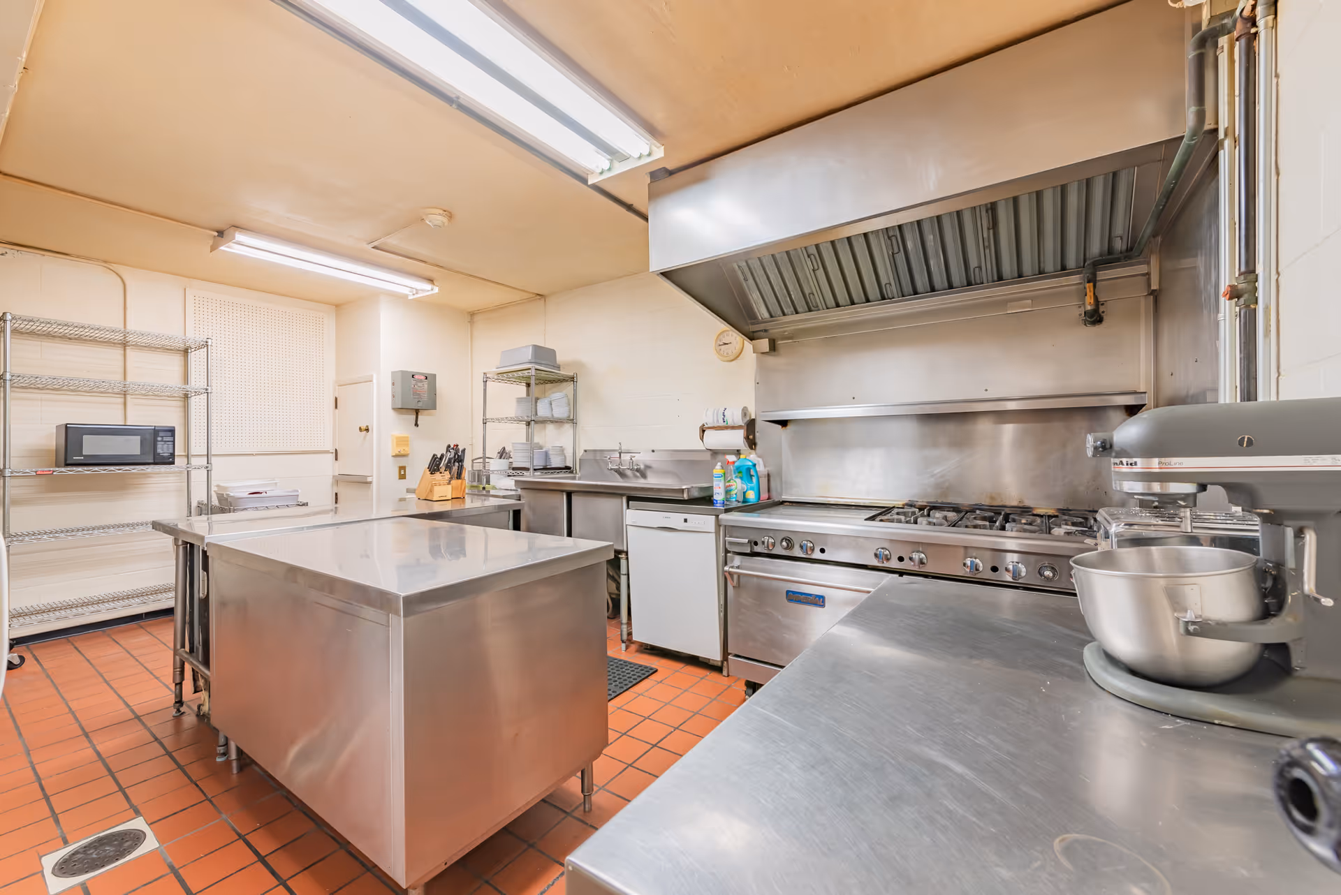 A commercial kitchen with stainless steel appliances and surfaces, including a large stove with an exhaust hood, a mixer, a microwave on a metal rack, and a central stainless steel island. The floor is covered with red tiles and the walls are light-colored.