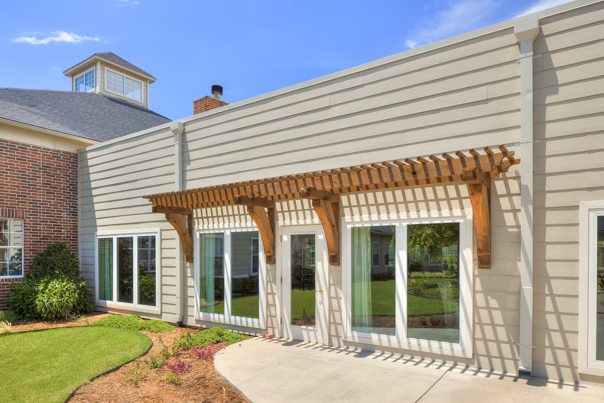 Exterior view of a senior living facility building with beige siding and a brick section. There are large windows and a wooden pergola casting shadows on the wall. The area in front has a concrete walkway and landscaped greenery with grass and small plants under a clear blue sky.