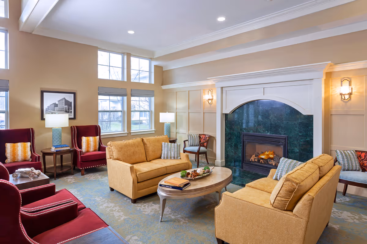 A cozy living room with two mustard yellow sofas facing each other around an oval wooden coffee table. The room features a green marble fireplace with a white mantel, two burgundy armchairs with patterned cushions, a side table with a lamp and books, and large windows letting in natural light. The walls are painted beige with decorative paneling and wall sconces providing additional lighting.