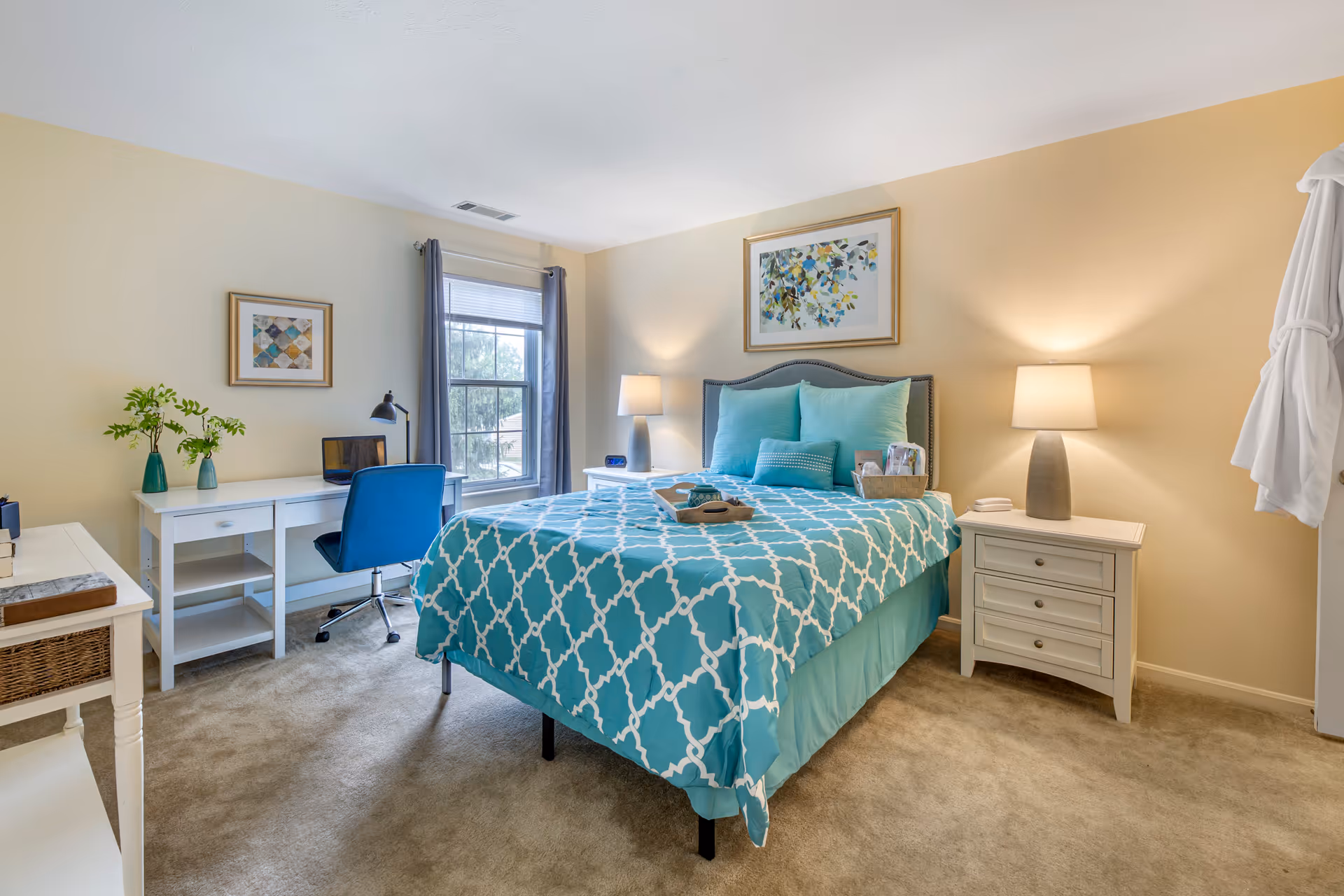 A bright and cozy bedroom with a bed covered in a turquoise and white patterned comforter, two matching pillows, and a small decorative pillow. The room has beige walls and carpeted floor. There are two white nightstands with lamps on either side of the bed. A framed artwork hangs above the bed. A window with gray curtains lets in natural light. A white desk with a blue chair and a small plant on top is positioned near the window. A white robe hangs on the wall to the right.