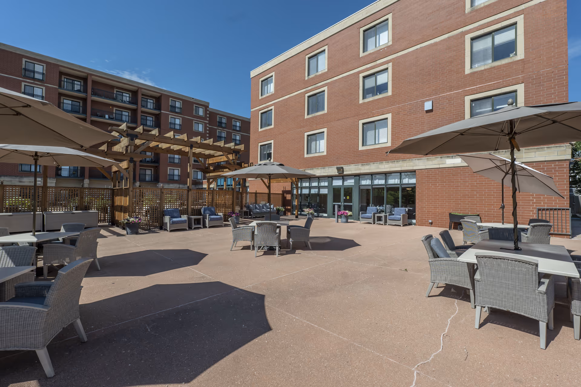 Outdoor courtyard patio with tables, umbrellas, lounge chairs and a wooden pergola beside a multi-story brick building.