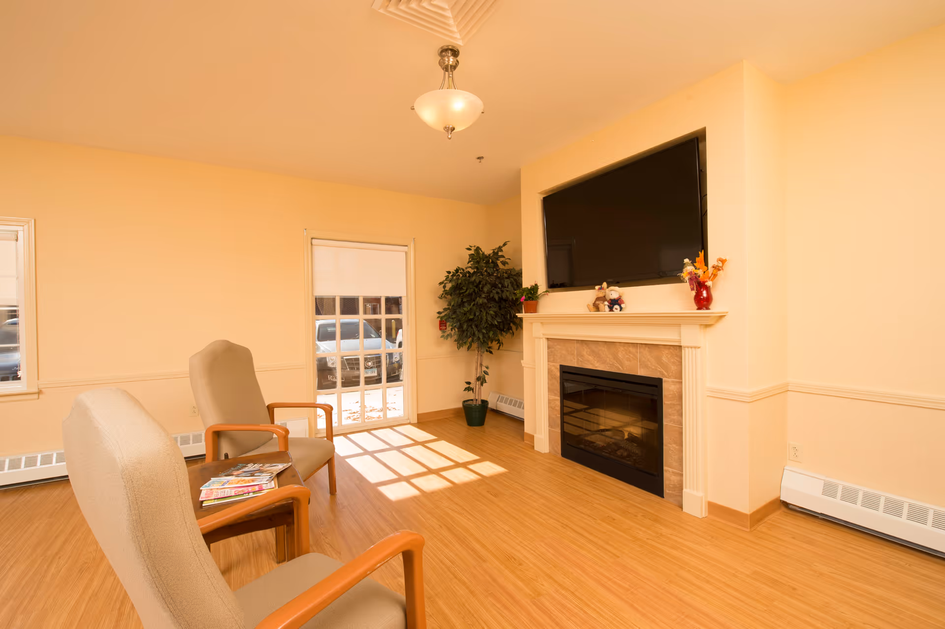 A cozy living room area with two beige armchairs and a small wooden table holding magazines. The room features a fireplace with a mounted flat-screen TV above it, a potted plant in the corner, and a window with a partially drawn blind letting in sunlight. The floor is wooden, and the walls are painted a warm beige color.