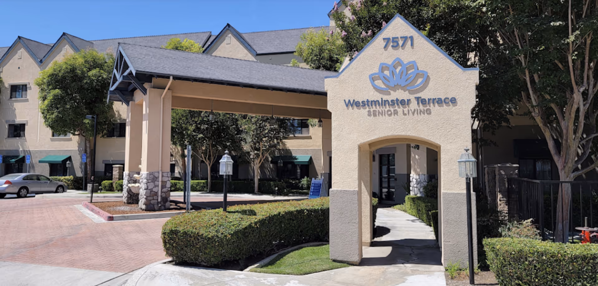 Exterior view of Westminster Terrace Senior Living building entrance with a covered driveway, beige walls, stone accents, green bushes, trees, and a clear blue sky.