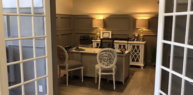 Small reception office with a central desk, two decorative chairs, lamps on a sideboard, and glass-pane French doors.