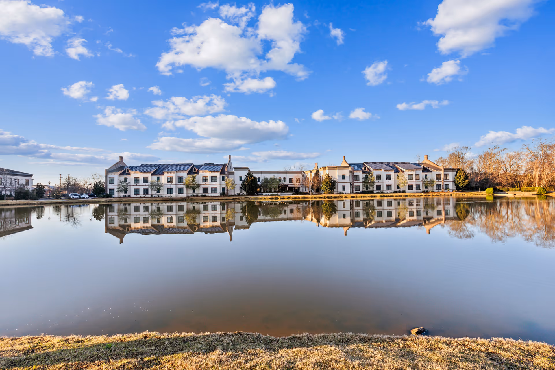 A large senior living facility named The Blake at Township is seen across a calm pond, reflecting the building and the blue sky with scattered clouds above. The building has a modern design with white walls and dark roofs, surrounded by some trees and grass.