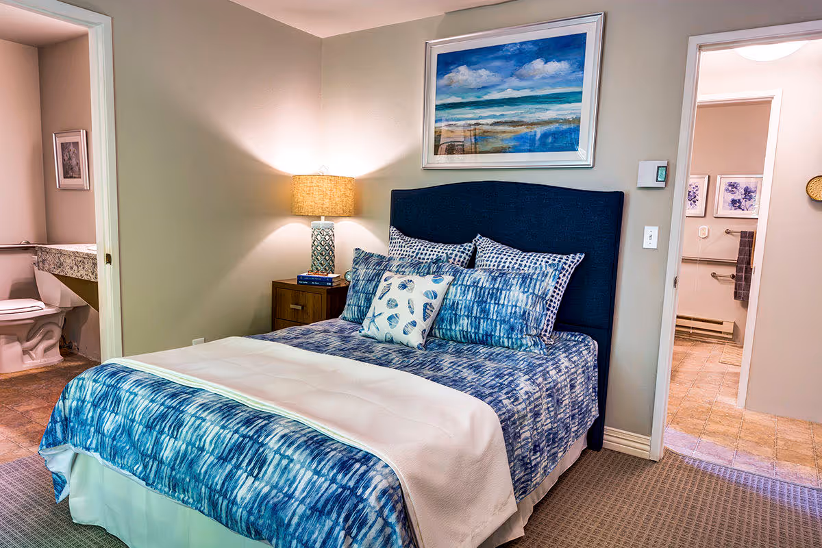 A bedroom with a blue upholstered headboard and blue patterned bedding. There are multiple pillows on the bed, including one with a seashell design. A wooden nightstand with a lamp and books is beside the bed. Two doorways are visible, one leading to a bathroom with a toilet and granite countertop, and the other to another room with tiled flooring and framed artwork on the walls. A large framed painting of a beach scene hangs above the bed.