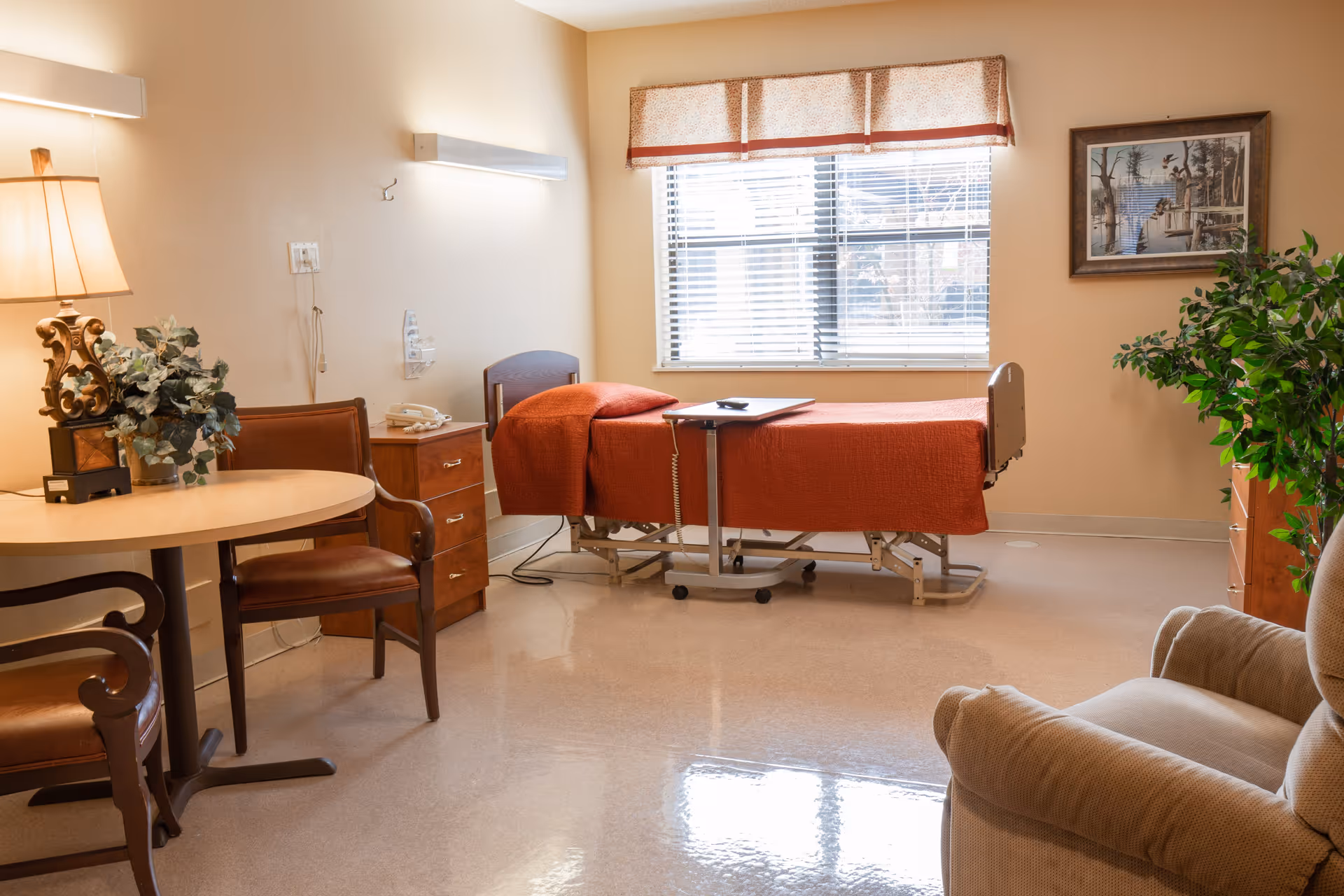 Sunlit nursing facility patient room with a single bed covered in an orange blanket, a small table and chairs, an armchair, and a window.