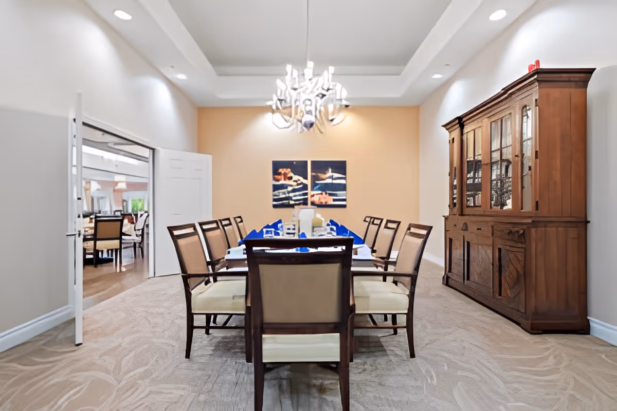 A formal dining room with a long rectangular table set with blue napkins and glassware. The room features beige walls, a large wooden cabinet on the right, a modern chandelier hanging from the ceiling, and an open doorway leading to another dining area.