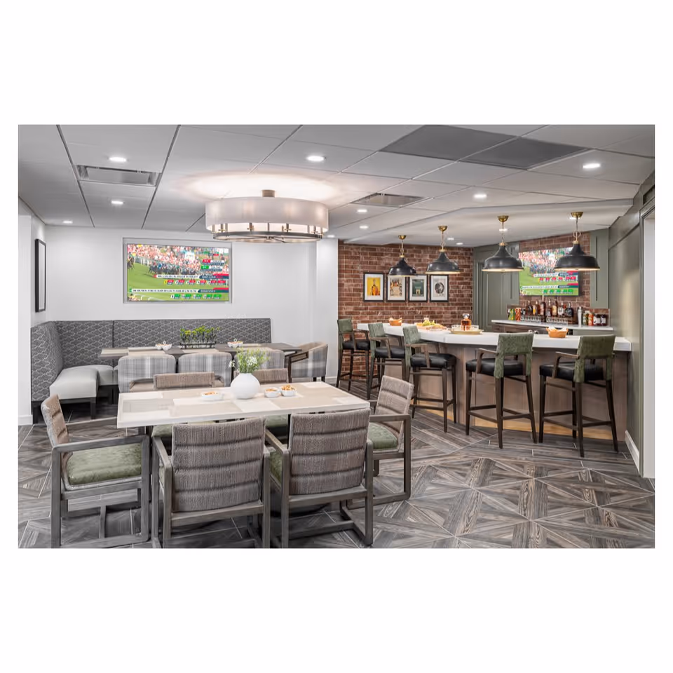 A modern dining and bar area with a large table surrounded by cushioned chairs in the foreground, a corner booth with a table and chairs along the left wall, and a bar with high stools on the right. The room features a patterned tile floor, a brick accent wall with framed artwork, two flat-screen TVs showing a sports event, and pendant lights hanging above the bar.