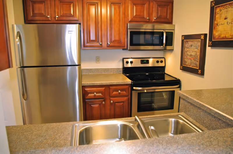 A kitchen with stainless steel appliances including a refrigerator, microwave, and electric stove. The kitchen has wooden cabinets, granite countertops, a double sink, and two framed chocolate-themed pictures on the wall.